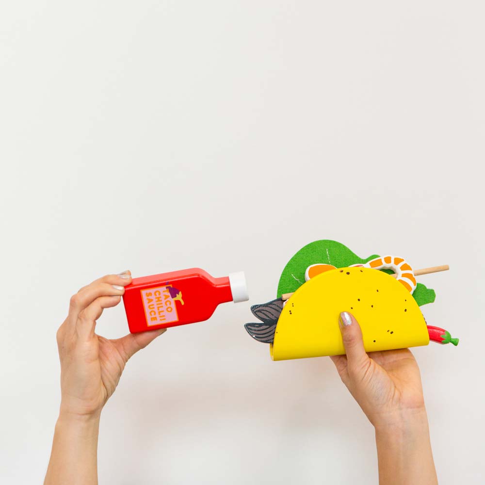 Make Me Iconic 21-piece wooden toy play Taco Kit featuring stand-up taco shells, prawns on skewers, paper takeaway bags, felt toppings, and pretend condiments like chilli sauce and sour cream for imaginative role play.
