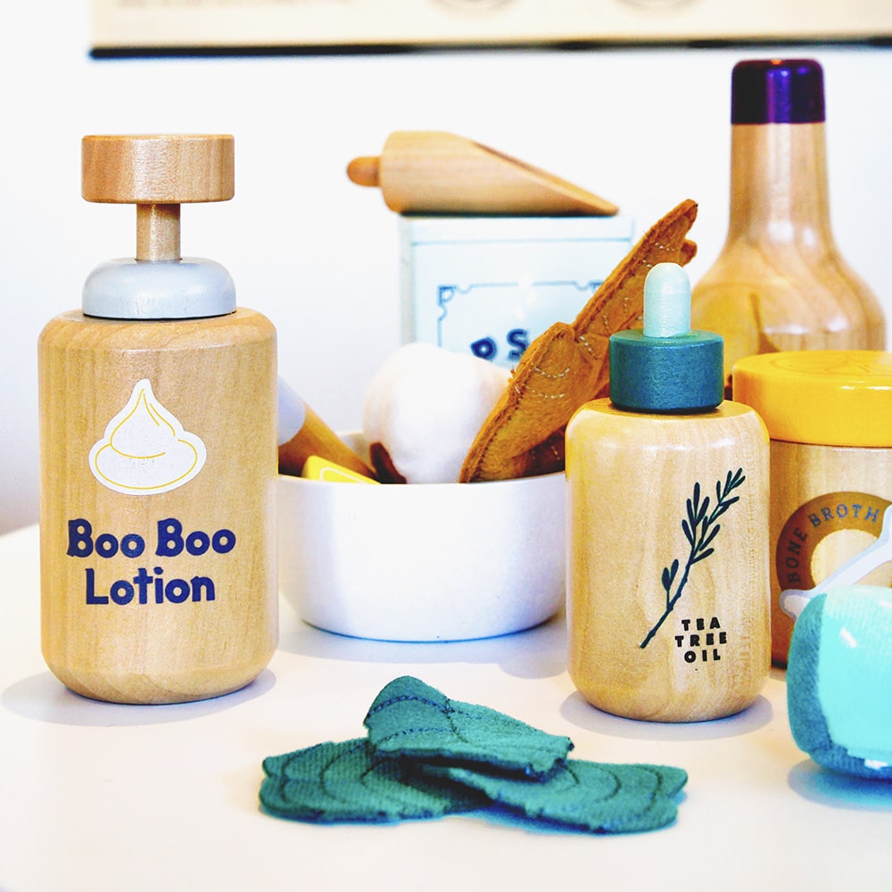 Wooden toy Natural Healers Kit with pestle and mortar, pretend Epsom salts, Manuka honey, bone broth, Tea Tree oil dropper, Boo Boo lotion, vitamins, arnica cream, herbal tea bags, and felt herbs for imaginative wellness play.