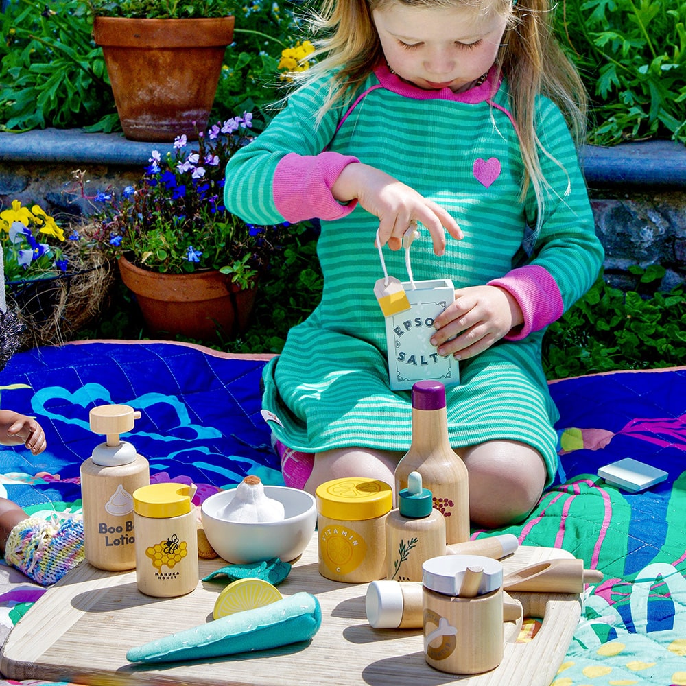 Wooden toy Natural Healers Kit with pestle and mortar, pretend Epsom salts, Manuka honey, bone broth, Tea Tree oil dropper, Boo Boo lotion, vitamins, arnica cream, herbal tea bags, and felt herbs for imaginative wellness play.
