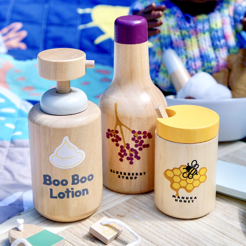 Wooden toy Natural Healers Kit with pestle and mortar, pretend Epsom salts, Manuka honey, bone broth, Tea Tree oil dropper, Boo Boo lotion, vitamins, arnica cream, herbal tea bags, and felt herbs for imaginative wellness play.