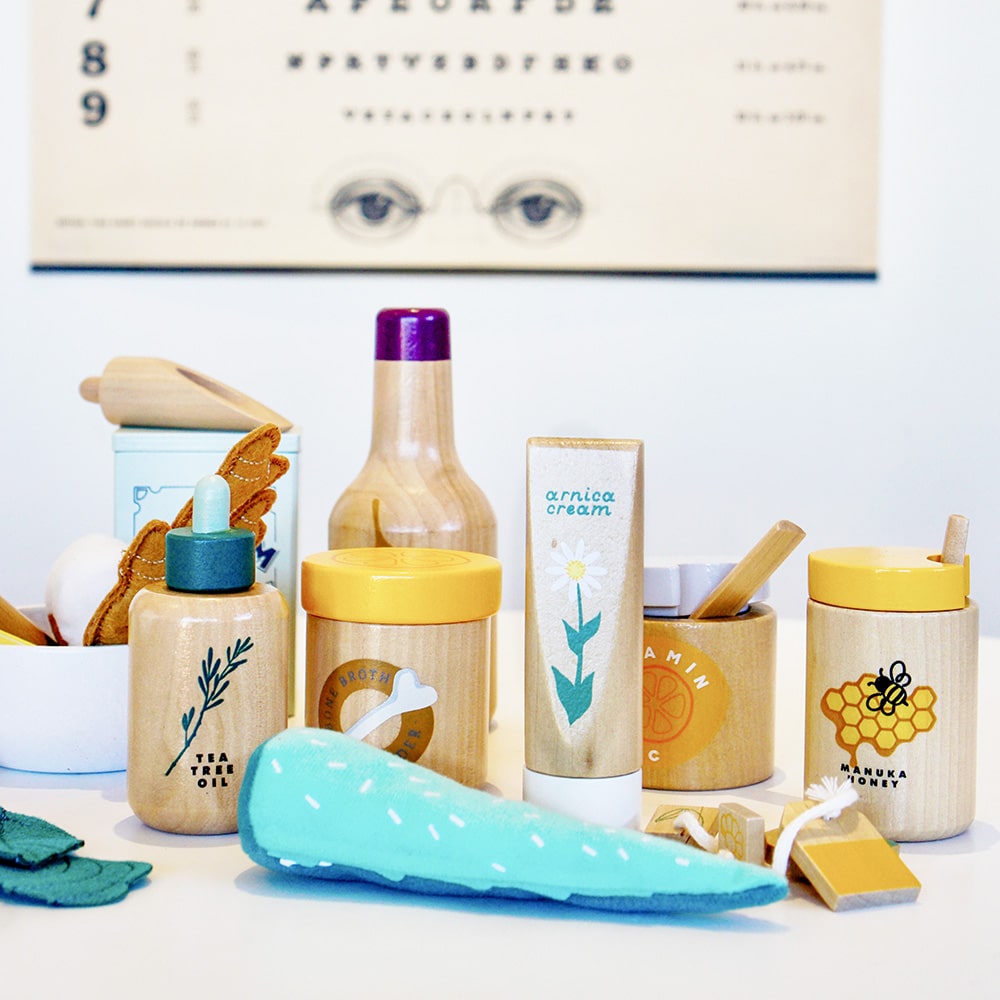 Wooden toy Natural Healers Kit with pestle and mortar, pretend Epsom salts, Manuka honey, bone broth, Tea Tree oil dropper, Boo Boo lotion, vitamins, arnica cream, herbal tea bags, and felt herbs for imaginative wellness play.