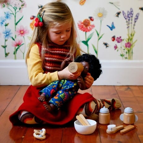 Child caring for a doll using a wooden doll accessory kit, brushing hair and feeding during nurturing open-ended play