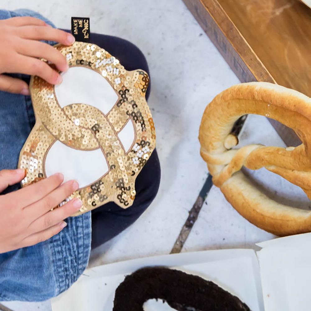 Make Me Iconic sequin pretzel purse with sparkling gold and pink sequins shaped like a pretzel, designed for holding small essentials like coins, keys, or hair clips. A fun, eye-catching accessory for kids, teens, and anyone who loves a playful touch of glamour.