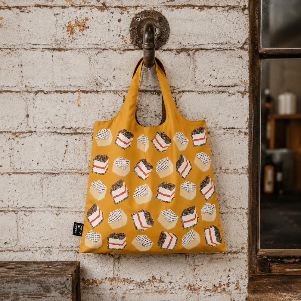 A Make Me Iconic reusable foldable shopping bag featuring a nostalgic Australian lamington design on a playful background. Made from 100% recycled fabric, this lightweight bag folds into its own matching pouch, making it perfect for shopping, travel, or everyday use while celebrating one of Australia’s most iconic sweet treats.