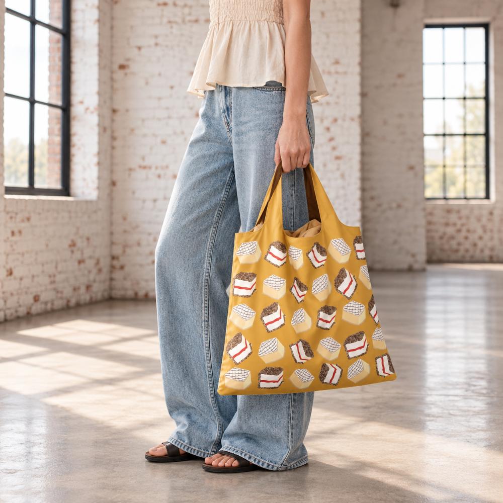 A Make Me Iconic reusable foldable shopping bag featuring a nostalgic Australian lamington design on a playful background. Made from 100% recycled fabric, this lightweight bag folds into its own matching pouch, making it perfect for shopping, travel, or everyday use while celebrating one of Australia’s most iconic sweet treats.