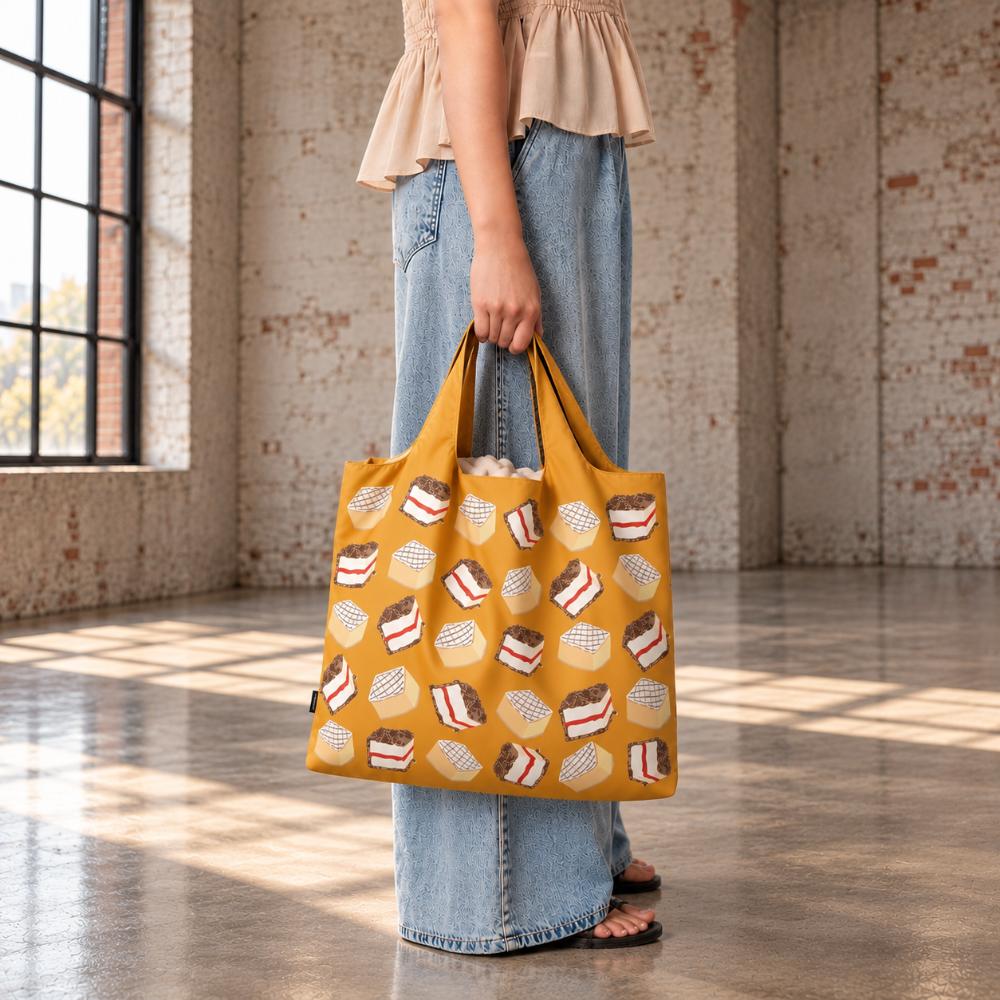 A Make Me Iconic reusable foldable shopping bag featuring a nostalgic Australian lamington design on a playful background. Made from 100% recycled fabric, this lightweight bag folds into its own matching pouch, making it perfect for shopping, travel, or everyday use while celebrating one of Australia’s most iconic sweet treats.