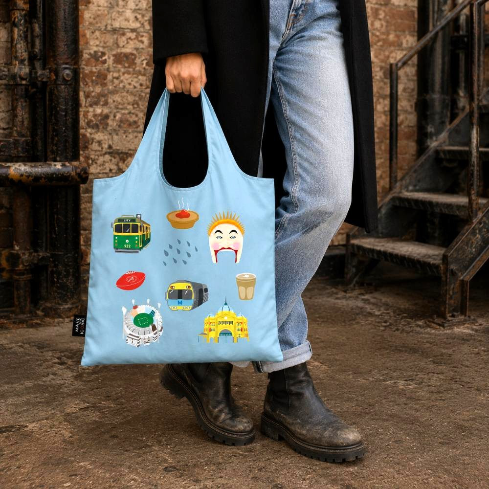 A Make Me Iconic reusable foldable shopping bag featuring iconic Melbourne landmarks and symbols including a W-Class tram, MCG, Flinders Street Station, coffee, meat pie, rain, and AFL football. Made from 100% recycled fabric, this lightweight eco-friendly tote folds into a compact pouch for shopping, travel, and everyday use.