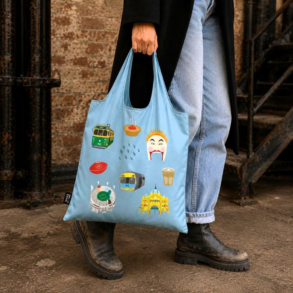 A Make Me Iconic reusable foldable shopping bag featuring iconic Melbourne landmarks and symbols including a W-Class tram, MCG, Flinders Street Station, coffee, meat pie, rain, and AFL football. Made from 100% recycled fabric, this lightweight eco-friendly tote folds into a compact pouch for shopping, travel, and everyday use.