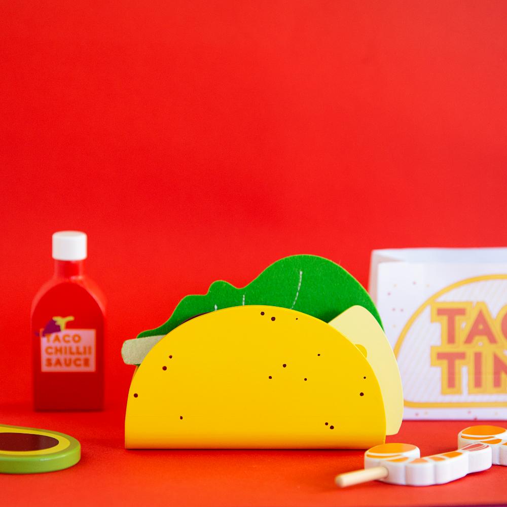 Make Me Iconic 21-piece wooden toy play Taco Kit featuring stand-up taco shells, prawns on skewers, paper takeaway bags, felt toppings, and pretend condiments like chilli sauce and sour cream for imaginative role play.