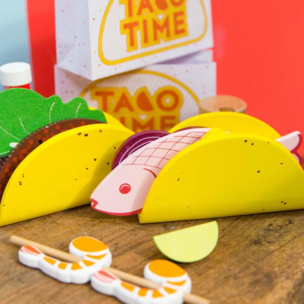 Make Me Iconic 21-piece wooden toy play Taco Kit featuring stand-up taco shells, prawns on skewers, paper takeaway bags, felt toppings, and pretend condiments like chilli sauce and sour cream for imaginative role play.