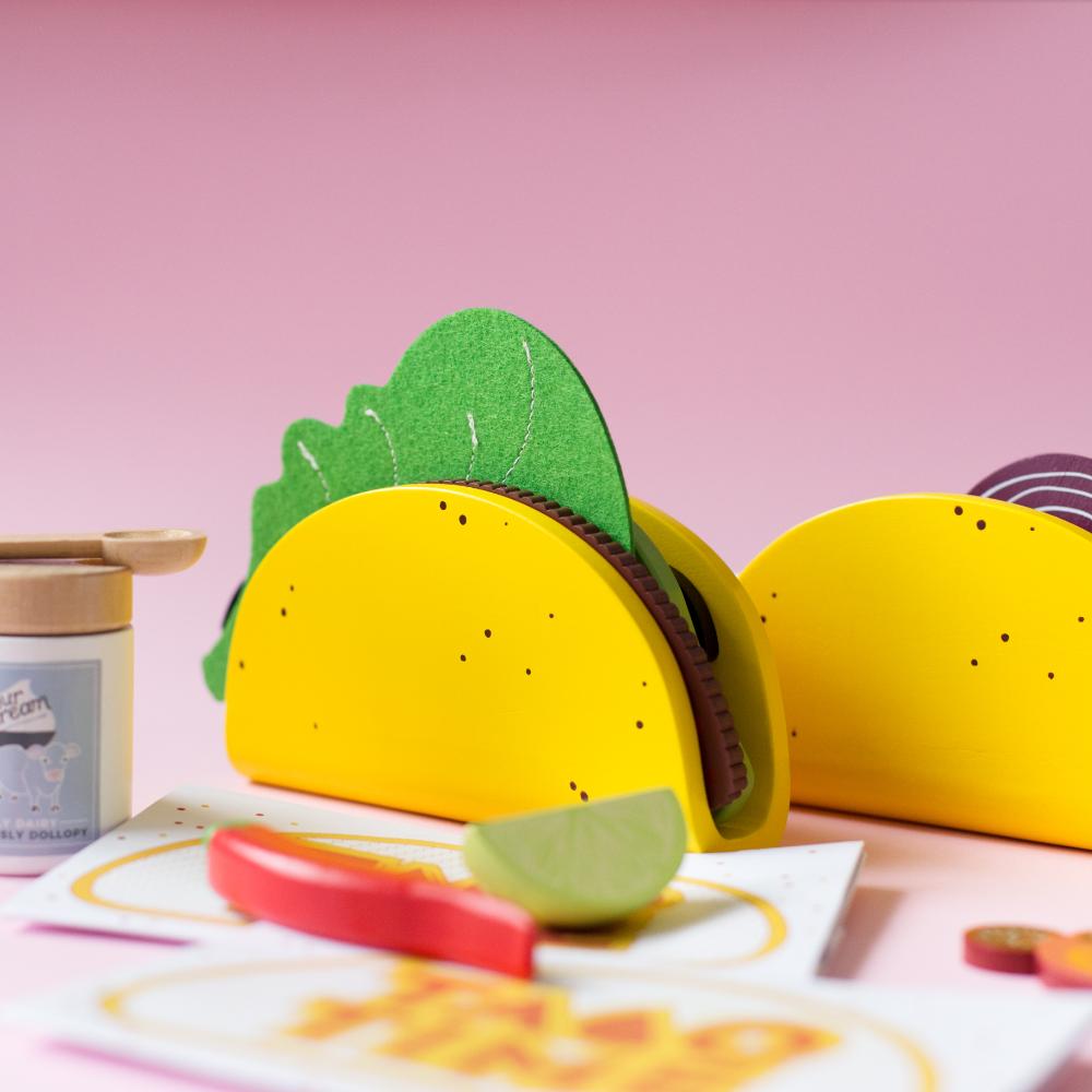 Make Me Iconic 21-piece wooden toy play Taco Kit featuring stand-up taco shells, prawns on skewers, paper takeaway bags, felt toppings, and pretend condiments like chilli sauce and sour cream for imaginative role play.