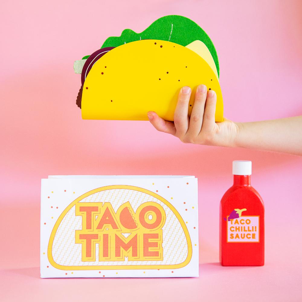 Make Me Iconic 21-piece wooden toy play Taco Kit featuring stand-up taco shells, prawns on skewers, paper takeaway bags, felt toppings, and pretend condiments like chilli sauce and sour cream for imaginative role play.