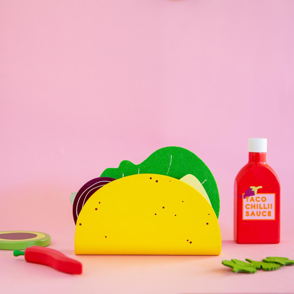 Make Me Iconic 21-piece wooden toy play Taco Kit featuring stand-up taco shells, prawns on skewers, paper takeaway bags, felt toppings, and pretend condiments like chilli sauce and sour cream for imaginative role play.