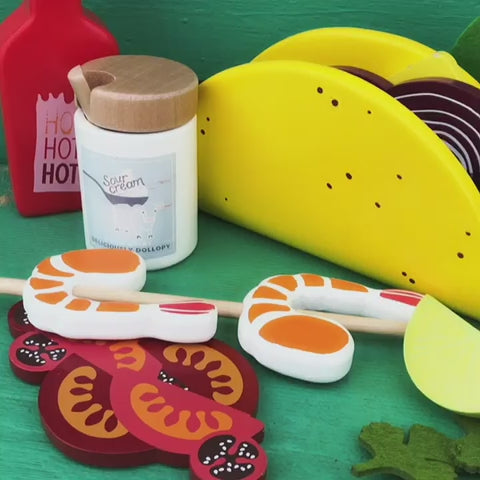Make Me Iconic 21-piece wooden toy play Taco Kit featuring stand-up taco shells, prawns on skewers, paper takeaway bags, felt toppings, and pretend condiments like chilli sauce and sour cream for imaginative role play.